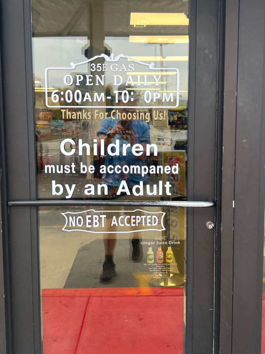 Glass door of a store displaying hours (6:00 AM–10:00 PM), a notice requiring children to be accompanied by an adult, and a sign stating “No EBT Accepted.” Store shelves and a red mat are visible inside.