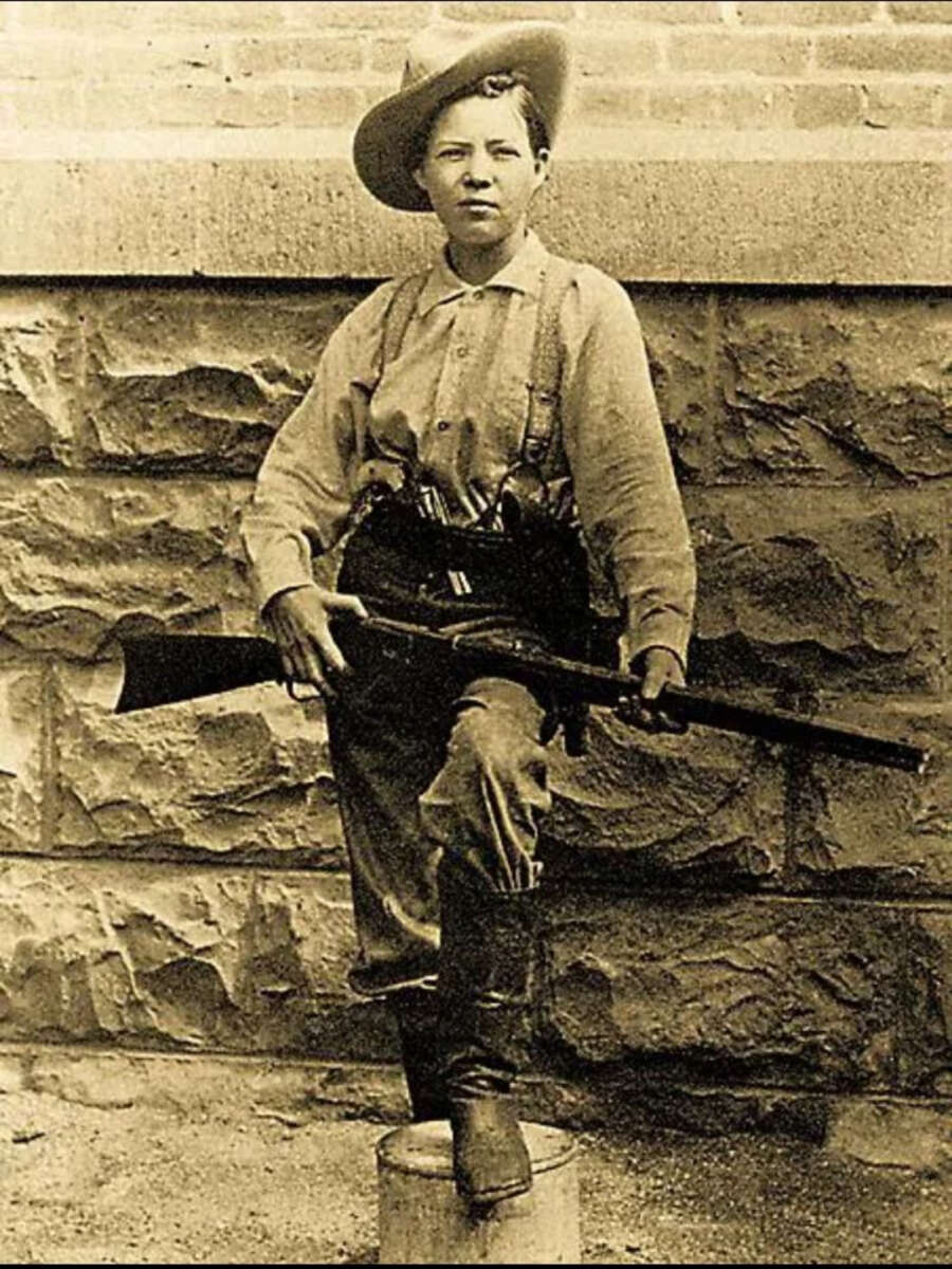 A sepia-toned historical photo of a young person in cowboy attire, wearing a wide-brimmed hat and boots, holding a rifle and resting one foot on a barrel, standing in front of a stone wall.