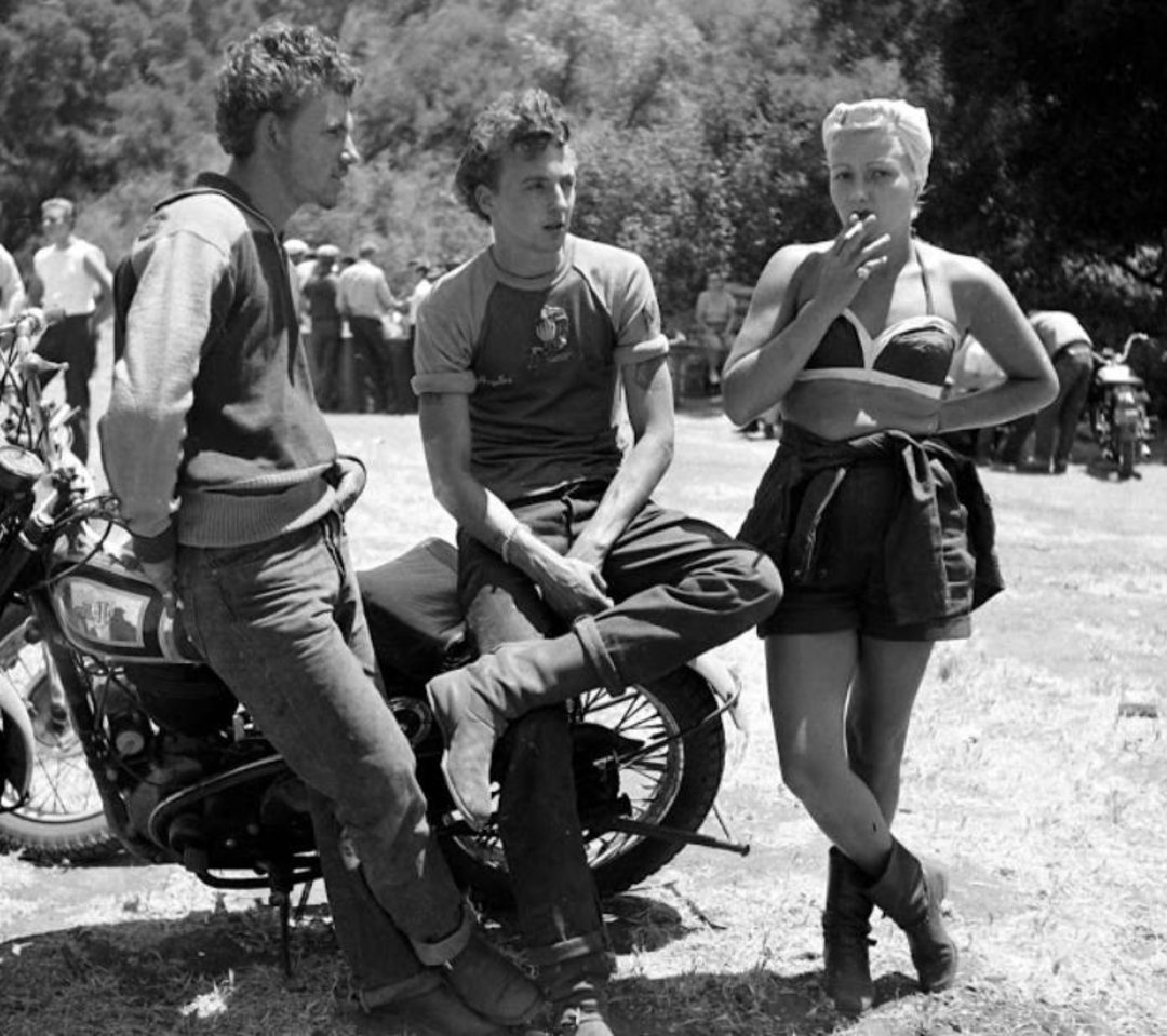 Three people in casual 1950s clothing gather outdoors near motorcycles. Two men stand and sit on a bike, while a woman in a bikini top and shorts stands, smoking a cigarette. More people and trees are visible in the background.