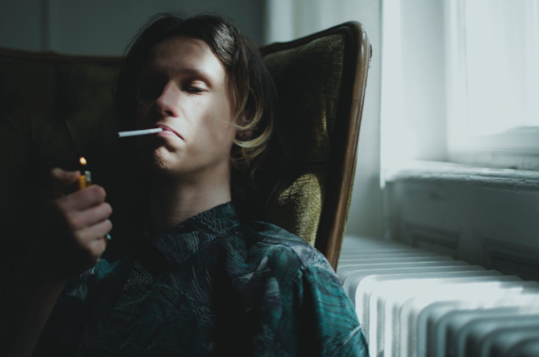 A person sits in a dimly lit room, holding a lighter to a cigarette in their mouth. Sunlight filters through a window, casting shadows on their face and the nearby radiator.
