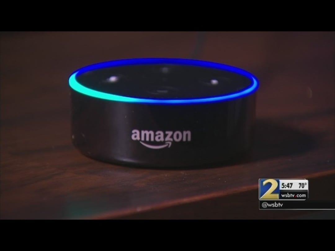 A close-up of an Amazon Echo Dot smart speaker with a glowing blue light ring, sitting on a wooden surface. A news station logo and time/weather graphic appear in the bottom right corner of the image.
