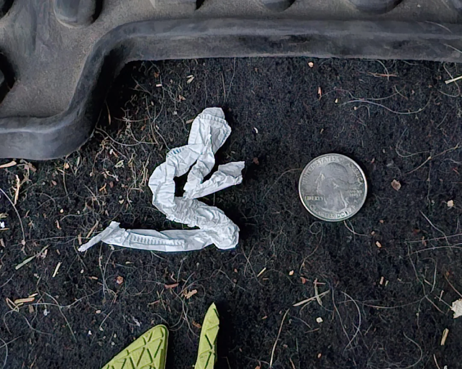 A crumpled white paper strip and a U.S. quarter lie on a dark, textured car floor mat, with part of a yellow shoe and a black rubber mat edge visible nearby.