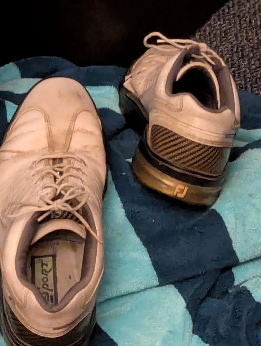 A pair of worn white athletic shoes with black soles resting on a blue and black checkered blanket. The shoes appear slightly dirty and are positioned at different angles.