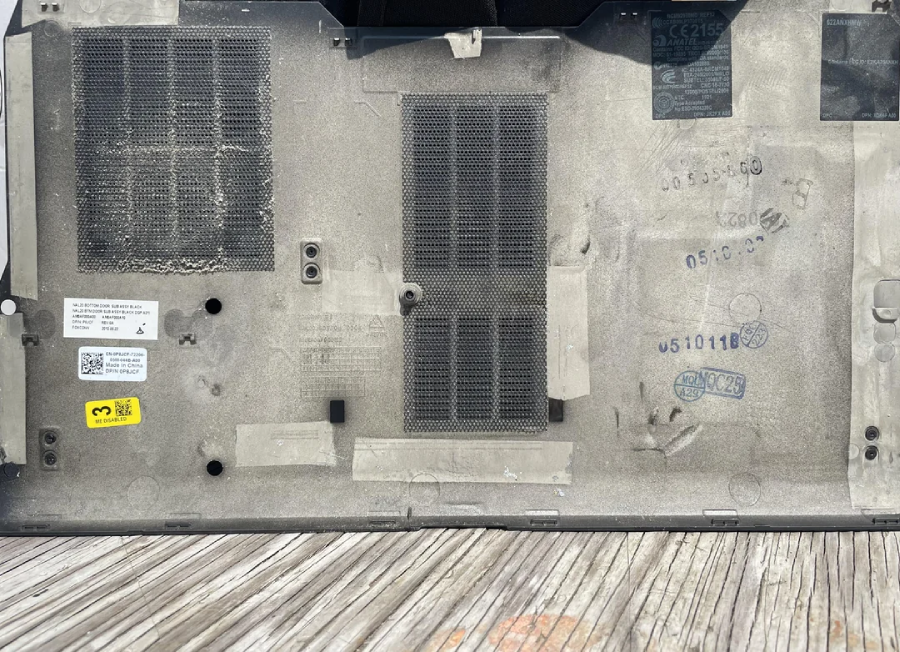 A close-up of a dusty, gray vented panel—likely the underside of a laptop or electronic device—showing mesh vents, stickers, printed numbers, and a textured wood surface below.