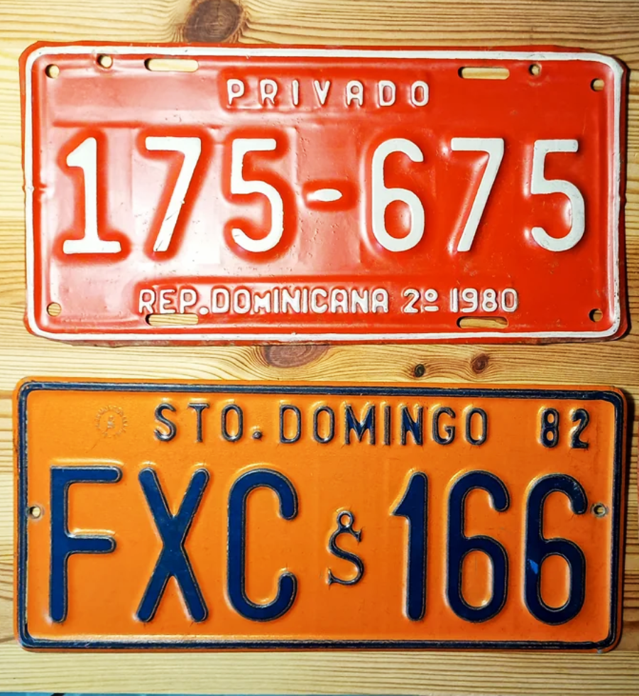 Two vintage Dominican Republic license plates, one red with white letters “175-675” and one orange with black letters “FXC § 166”, displayed on a wooden surface. Both show years: 1980 and 1982.