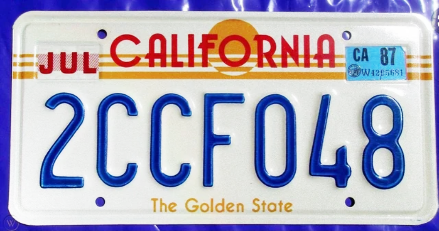 A California license plate with the number 2CCF048, featuring a red "CALIFORNIA" text, a yellow sunset stripe, “The Golden State” slogan, and registration stickers for July and the year 1987.