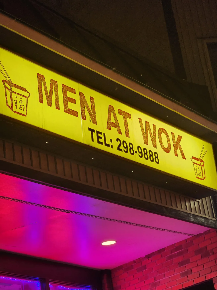 A yellow restaurant sign reads "MEN AT WOK" with a phone number "TEL: 298-9888" and images of takeout boxes with chopsticks on each side, photographed at night above a brick wall.