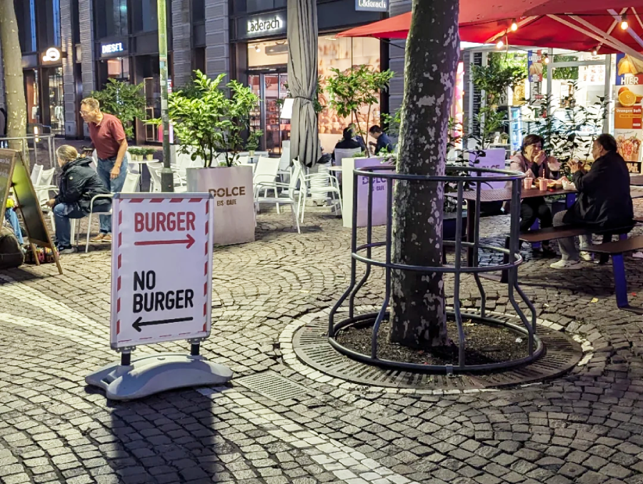 A sidewalk sign with arrows points left for "BURGER" and right for "NO BURGER" on a cobblestone plaza with outdoor seating. People are sitting at tables under red umbrellas and surrounded by plants.