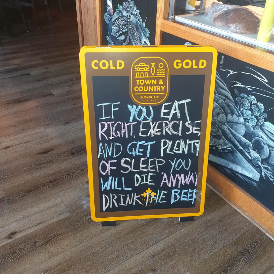 A chalkboard sign in a restaurant reads: "If you eat right, exercise and get plenty of sleep you will die anyway. Drink the beer." The sign is on a wooden floor next to a counter with bread and chalk art.
