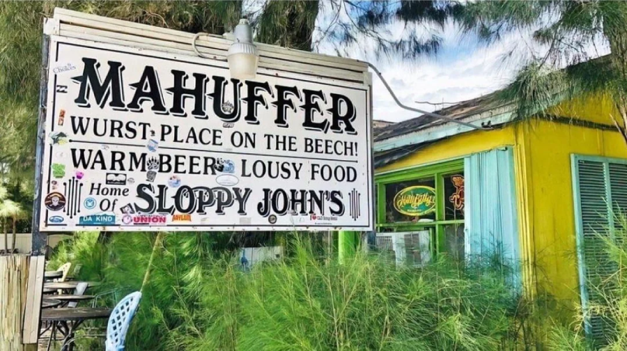 A large sign outside a rustic yellow building reads, “MAHUFFER: Wurst Place on the Beach! Warm Beer, Lousy Food, Home of Sloppy John’s,” surrounded by trees and greenery.