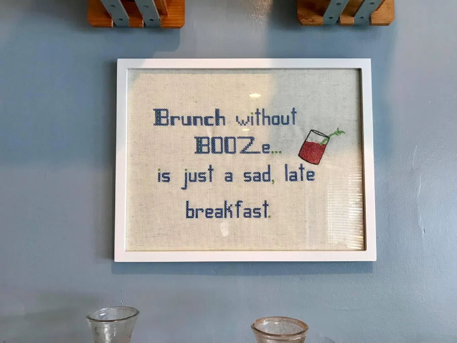 A framed cross-stitch hangs on a blue wall, reading “Brunch without BOOZe... is just a sad, late breakfast,” with a small stitched image of a cocktail glass beside the text.