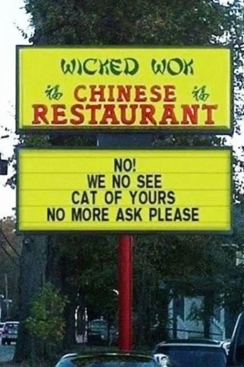 A restaurant sign reads: "WICKED WOK CHINESE RESTAURANT. NO! WE NO SEE CAT OF YOURS NO MORE ASK PLEASE." Trees and a street are visible in the background.