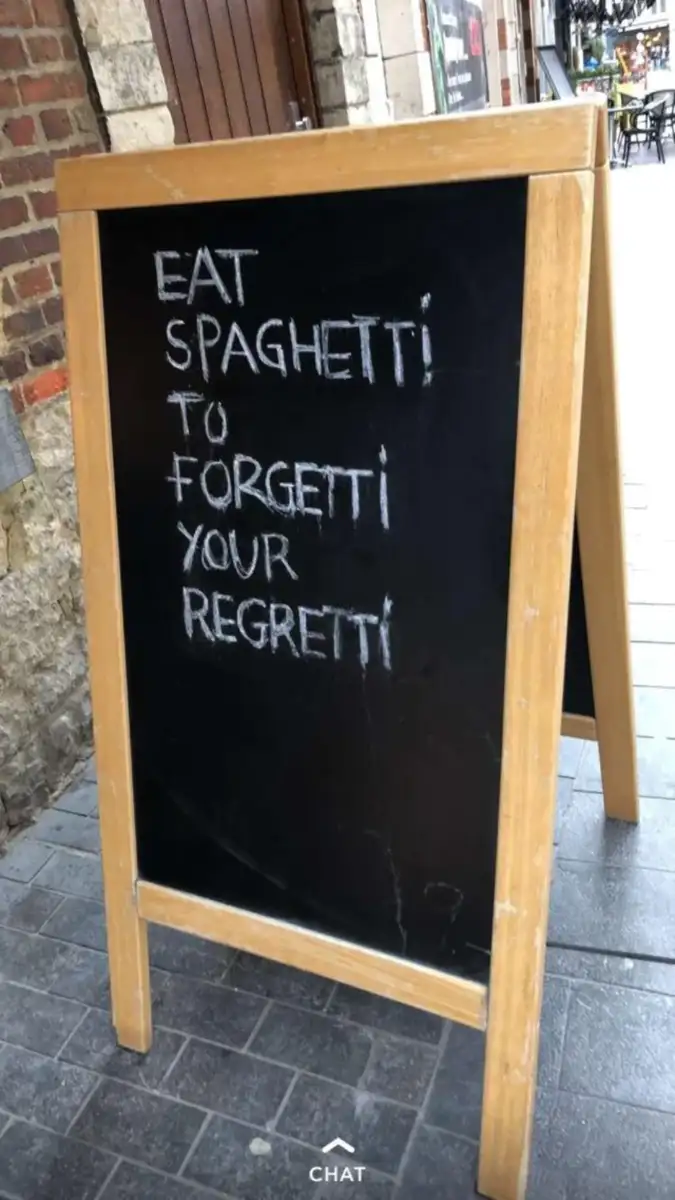 A sidewalk chalkboard sign reads, "EAT SPAGHETTI TO FORGETTI YOUR REGRETTI," written in uneven white chalk on a black board with a wooden frame.