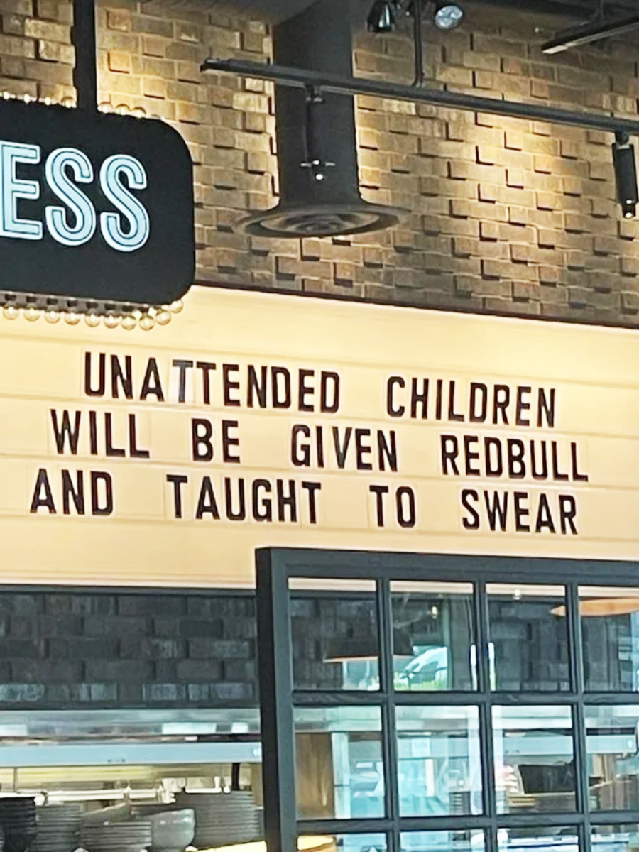 A sign in a restaurant reads: "UNATTENDED CHILDREN WILL BE GIVEN REDBULL AND TAUGHT TO SWEAR," using black letters on a light background, with brick walls and shelves visible below.