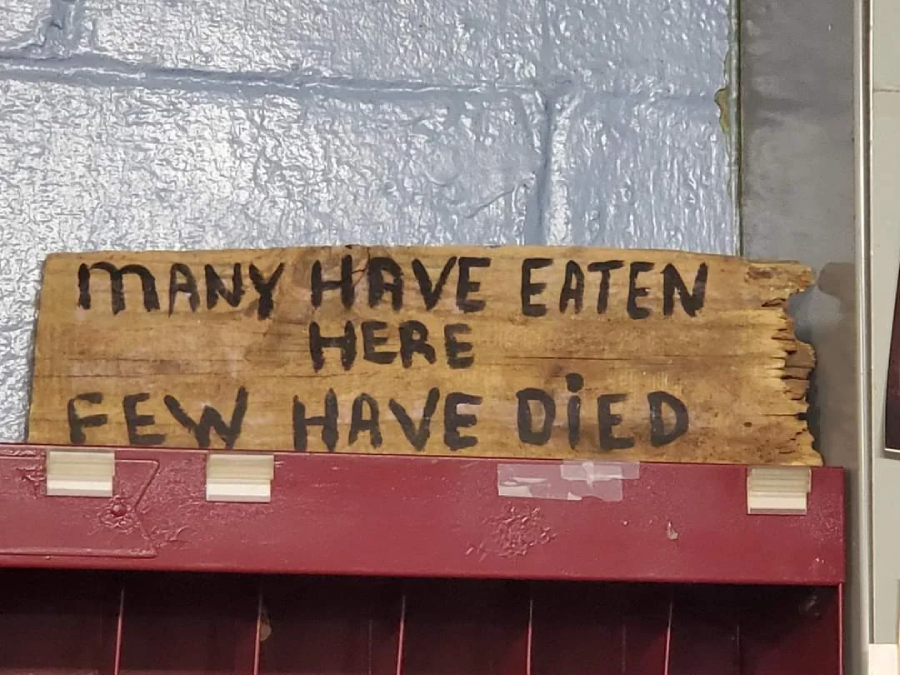 A weathered wooden sign with black, uneven letters reads: "MANY HAVE EATEN HERE FEW HAVE DIED." It sits on a red shelf against a blue-gray textured wall.
