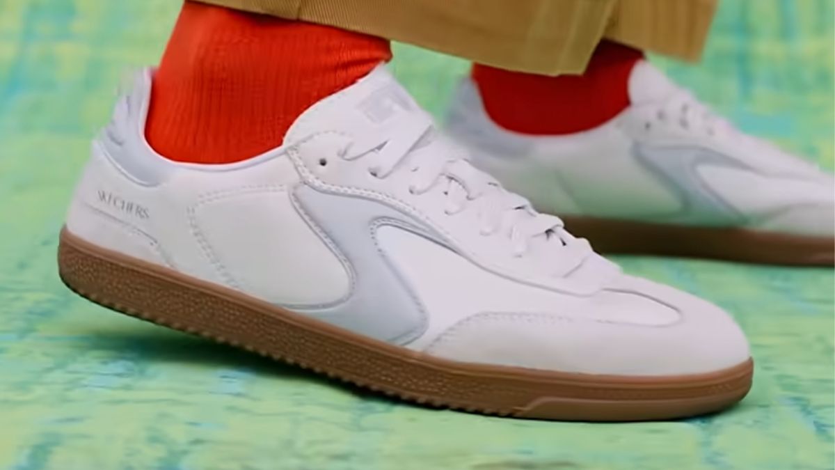 A person wearing white sneakers with a gum sole and red socks, standing on a blue-green surface.