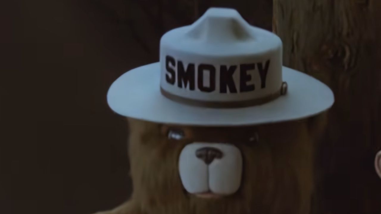 A close-up of a bear mascot wearing a white ranger hat labeled “SMOKEY,” standing in front of a tree.