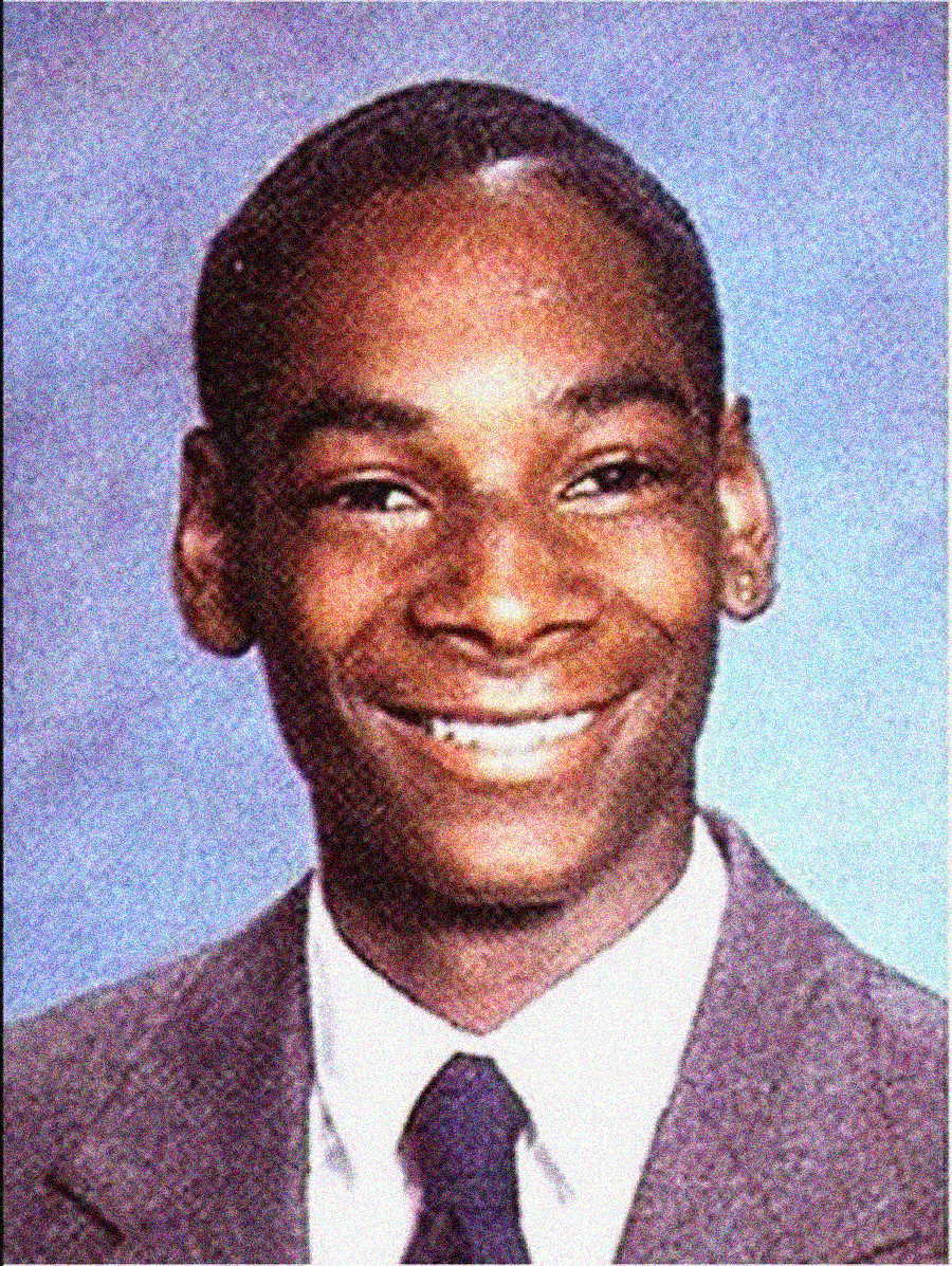 A young man wearing a suit and tie smiles at the camera in a formal yearbook-style portrait with a blue background.