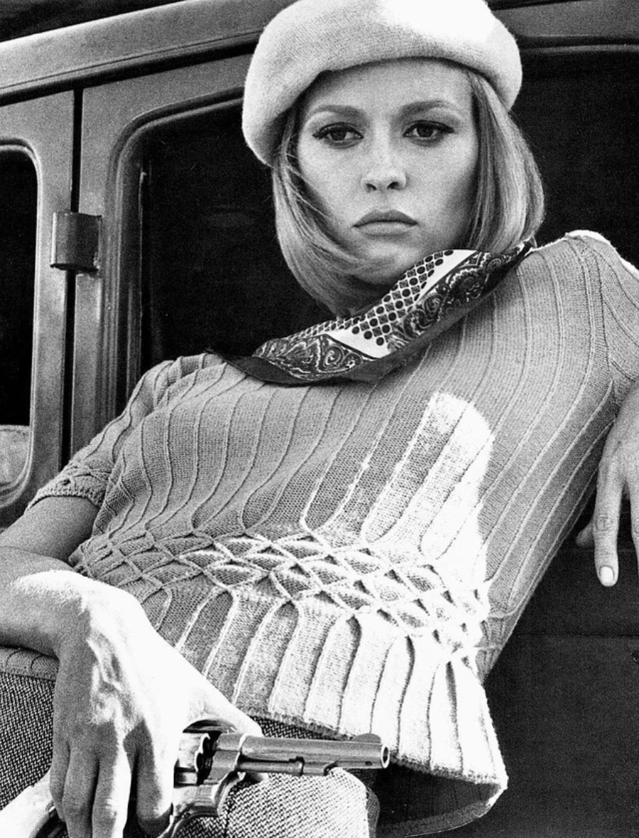 Black-and-white photo of a woman in a beret and patterned scarf, leaning against a car, holding a revolver with a serious expression.