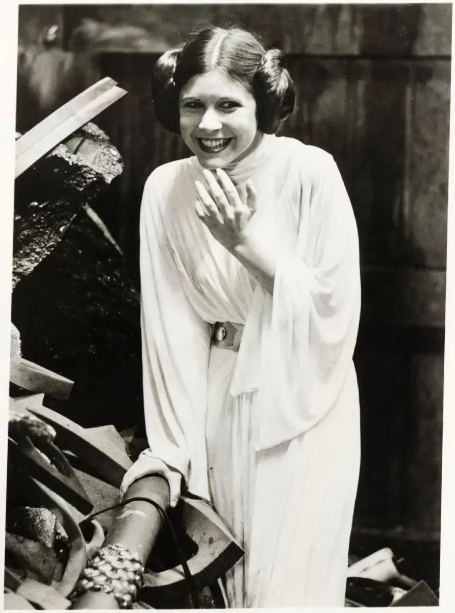 A woman dressed in a white gown with long sleeves and a belt, wearing her hair in two side buns, stands among debris and laughs, covering her mouth with one hand.