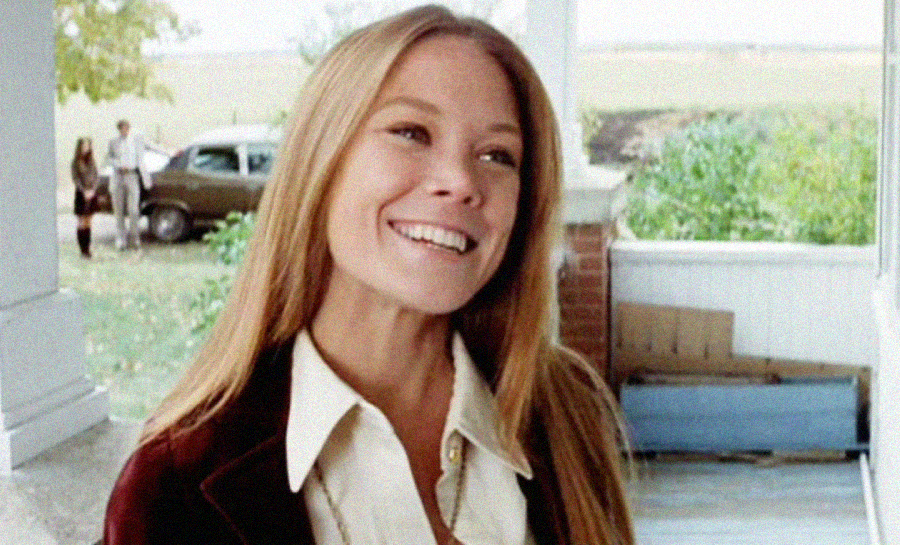 A smiling woman with long blonde hair stands outside on a porch. In the background, a man and a car are visible near some trees and open fields. The woman is wearing a cream-colored shirt and a dark jacket.