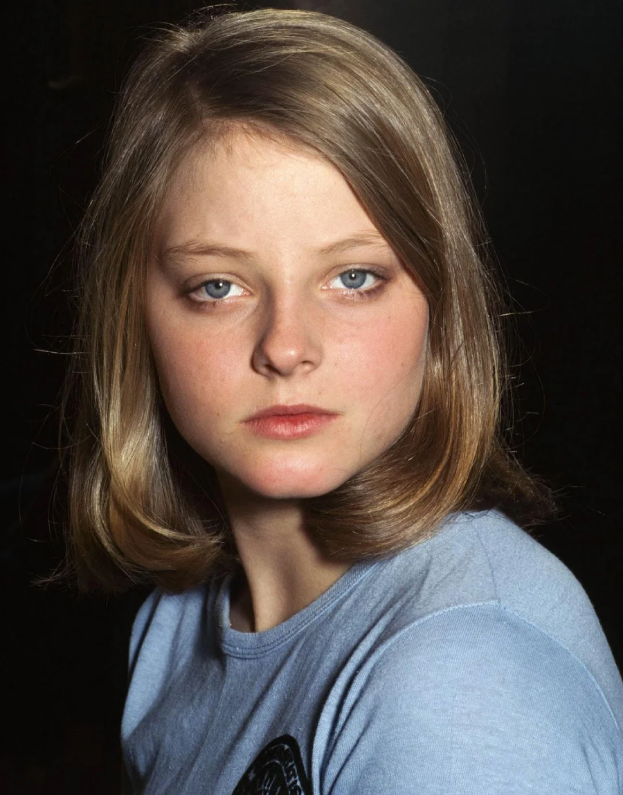 A young person with blonde hair and blue eyes wearing a light blue shirt, looking directly at the camera with a neutral expression against a dark background.