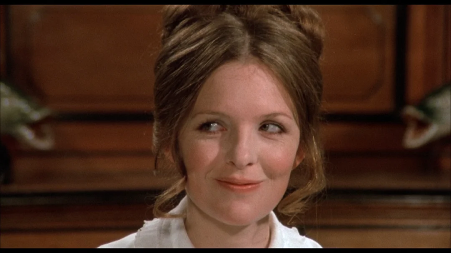 A woman with light skin and brown hair styled up is smirking and looking to her left. She wears a white top and sits in front of a wooden background.