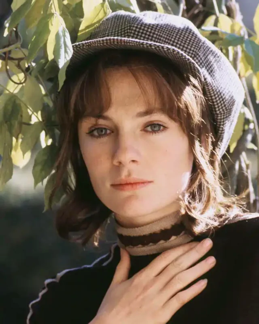 A young woman with blue eyes and brown hair wears a checkered cap and a striped turtleneck, standing outdoors near green leaves. She gazes calmly at the camera with one hand resting on her neck.