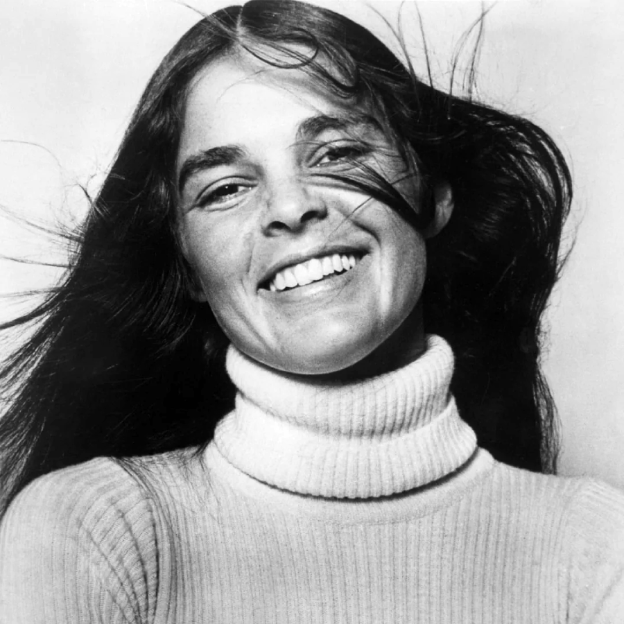 Black and white photo of a woman smiling with long dark hair blown back, wearing a ribbed turtleneck sweater. The background is plain and light-colored.