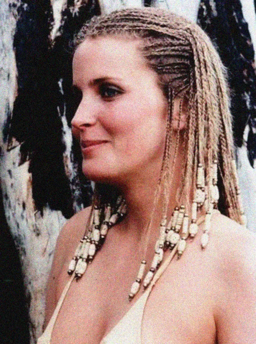 A woman with light skin and braided blonde hair decorated with white beads stands outdoors, wearing a light-colored swimsuit top. The background features a tree with dark and light bark.
