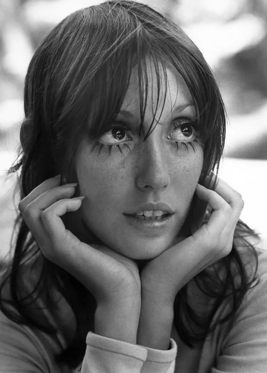 Black-and-white photo of a woman with long hair, resting her chin on her hands. She has large, expressive eyes with long lashes and slightly parted lips, gazing upward with a thoughtful expression.