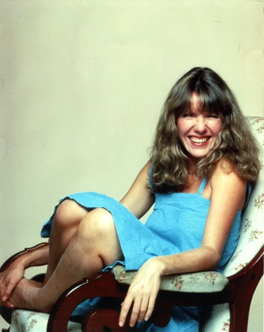 A woman with long, wavy hair in a blue dress sits cross-legged on a floral armchair, smiling widely and looking at the camera against a plain light background.