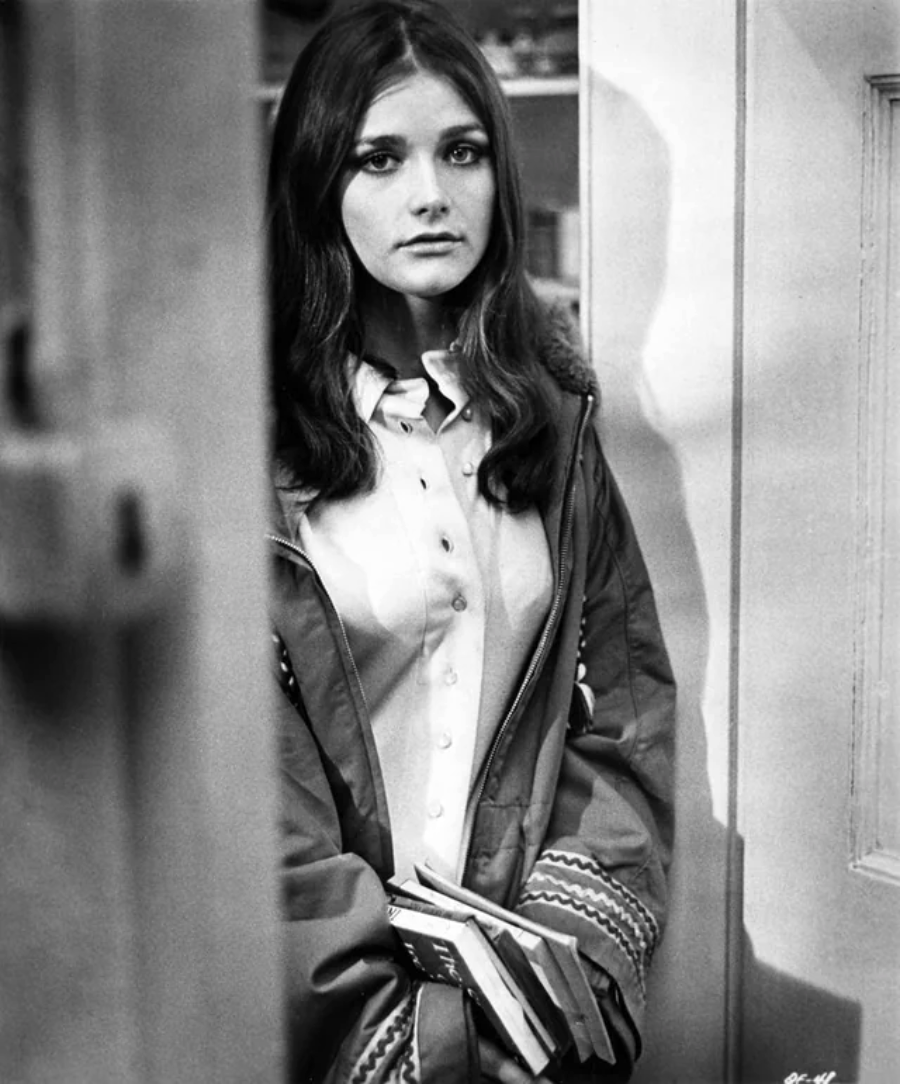 A young woman with long dark hair stands indoors, wearing a buttoned shirt and a jacket. She looks directly at the camera with a neutral expression and holds books in her hands. The background is partially obscured by a door.