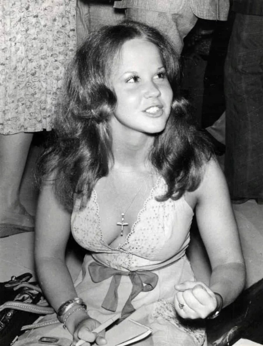 A young woman with long hair sits on the floor, wearing a light-colored dress with a bow and a cross necklace. She is looking slightly upward and appears to be in conversation, with people standing around her.