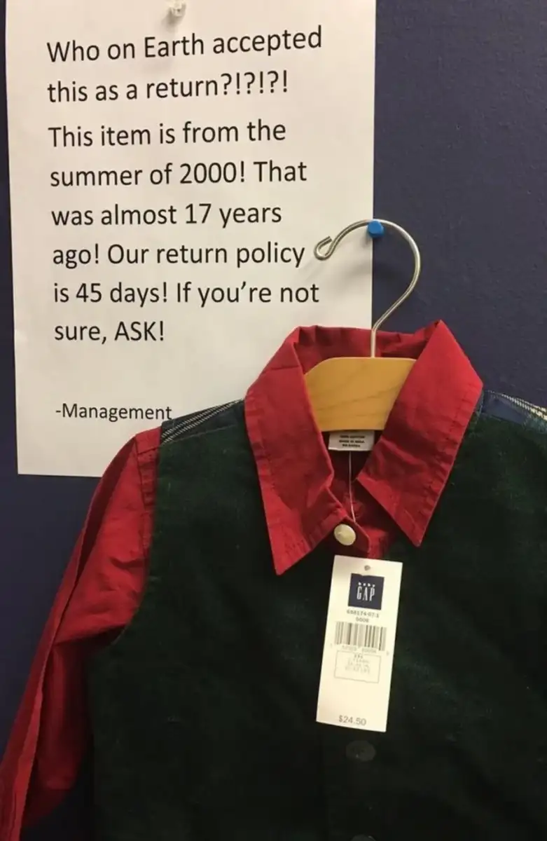 A red shirt and green vest on a hanger with a price tag, placed in front of a sign expressing frustration about someone accepting a return from the year 2000, despite the store’s 45-day return policy.