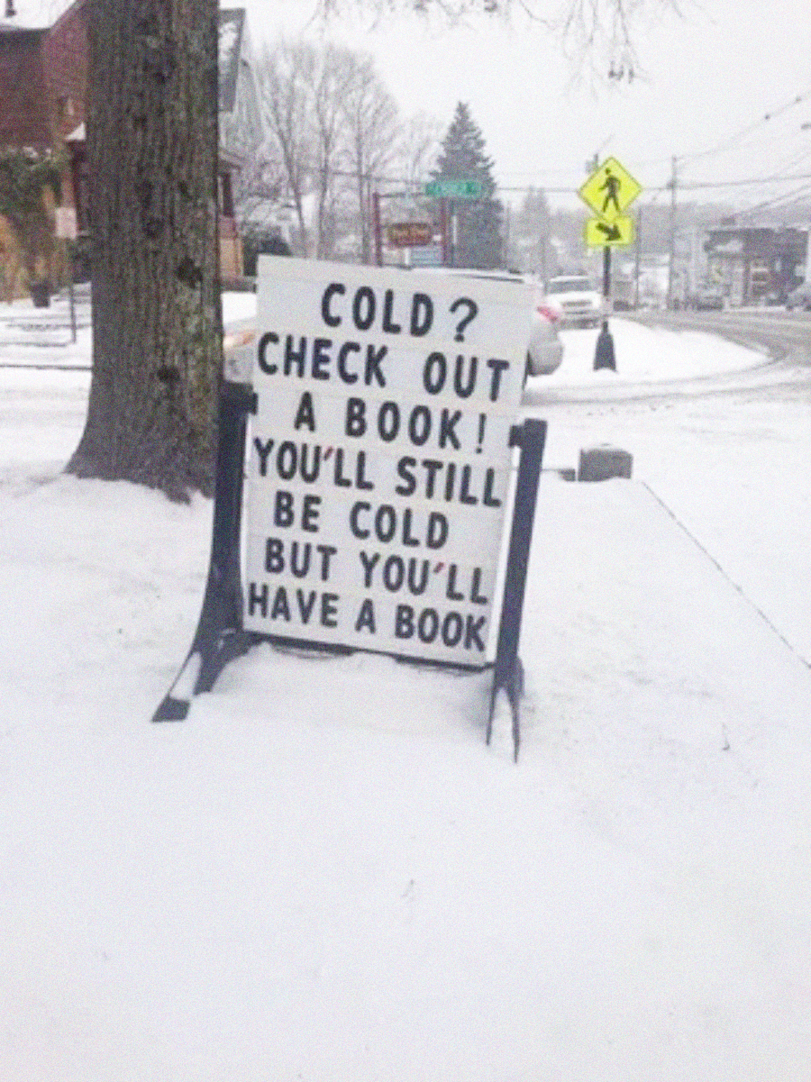 A sidewalk sign in a snowy street reads, "COLD? CHECK OUT A BOOK! YOU’LL STILL BE COLD BUT YOU’LL HAVE A BOOK." Snow covers the ground and trees; buildings and traffic signs are in the background.