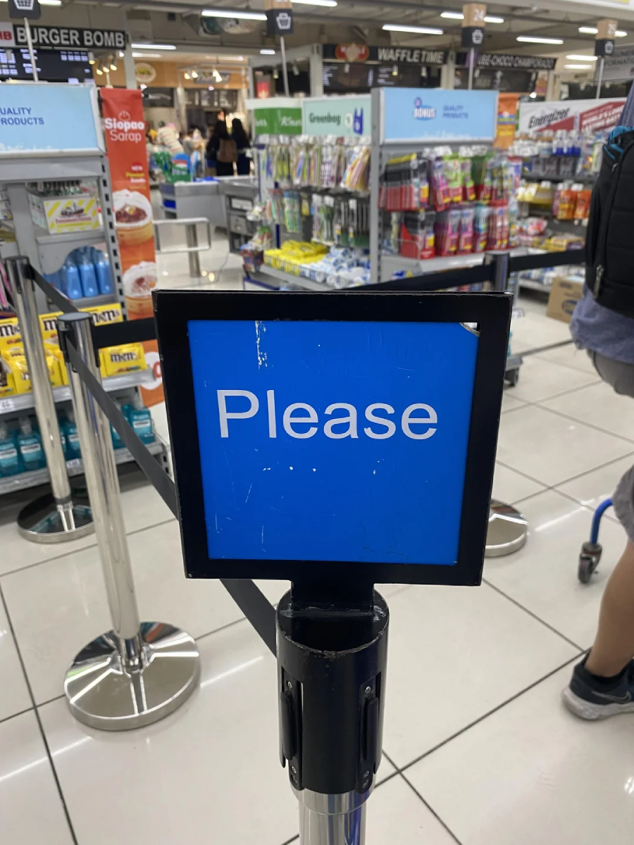 A blue sign in a supermarket says "Please," with checkout counters, store shelves, and a shopping cart visible in the background.