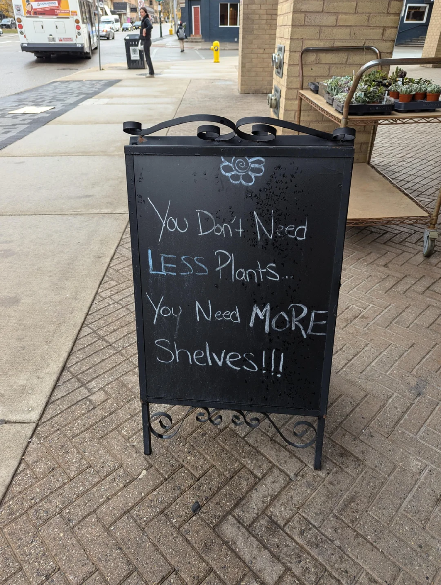 A chalkboard sign on a sidewalk reads: "You don't need LESS plants... You need MORE shelves!!!" with a small flower drawing above the text.