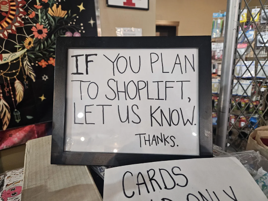 A framed sign reads, "IF YOU PLAN TO SHOPLIFT, LET US KNOW. Thanks." It sits on a counter in a store, with shelves of merchandise and a tapestry in the background.