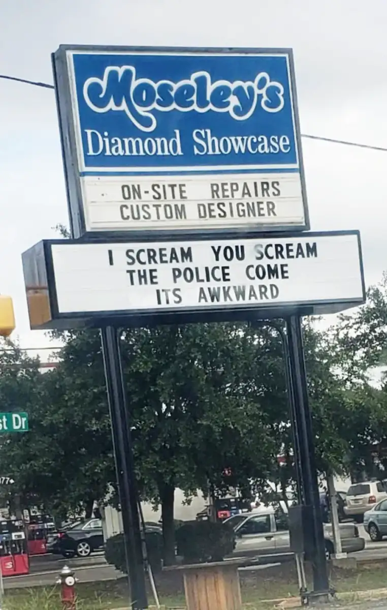 A jewelry store sign reads: "I SCREAM YOU SCREAM THE POLICE COME ITS AWKWARD." Above, the store name "Moseley's Diamond Showcase" is visible, along with "ON-SITE REPAIRS CUSTOM DESIGNER." Trees and a street are in the background.