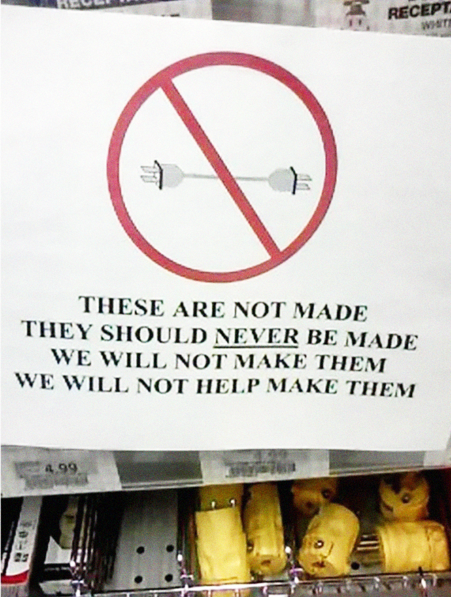 A sign with an illustration of a plug and outlet crossed out reads: “These are not made. They should never be made. We will not make them. We will not help make them.” Electrical plug adapters are visible below the sign.