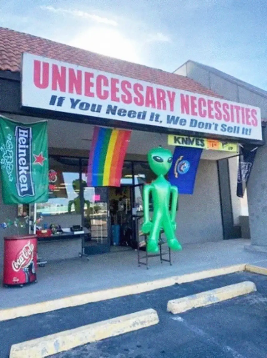 A storefront with a sign reading "UNNECESSARY NECESSITIES: If You Need It, We Don't Sell It!" has an inflatable green alien, a rainbow flag, and banners for Heineken and knives displayed outside.
