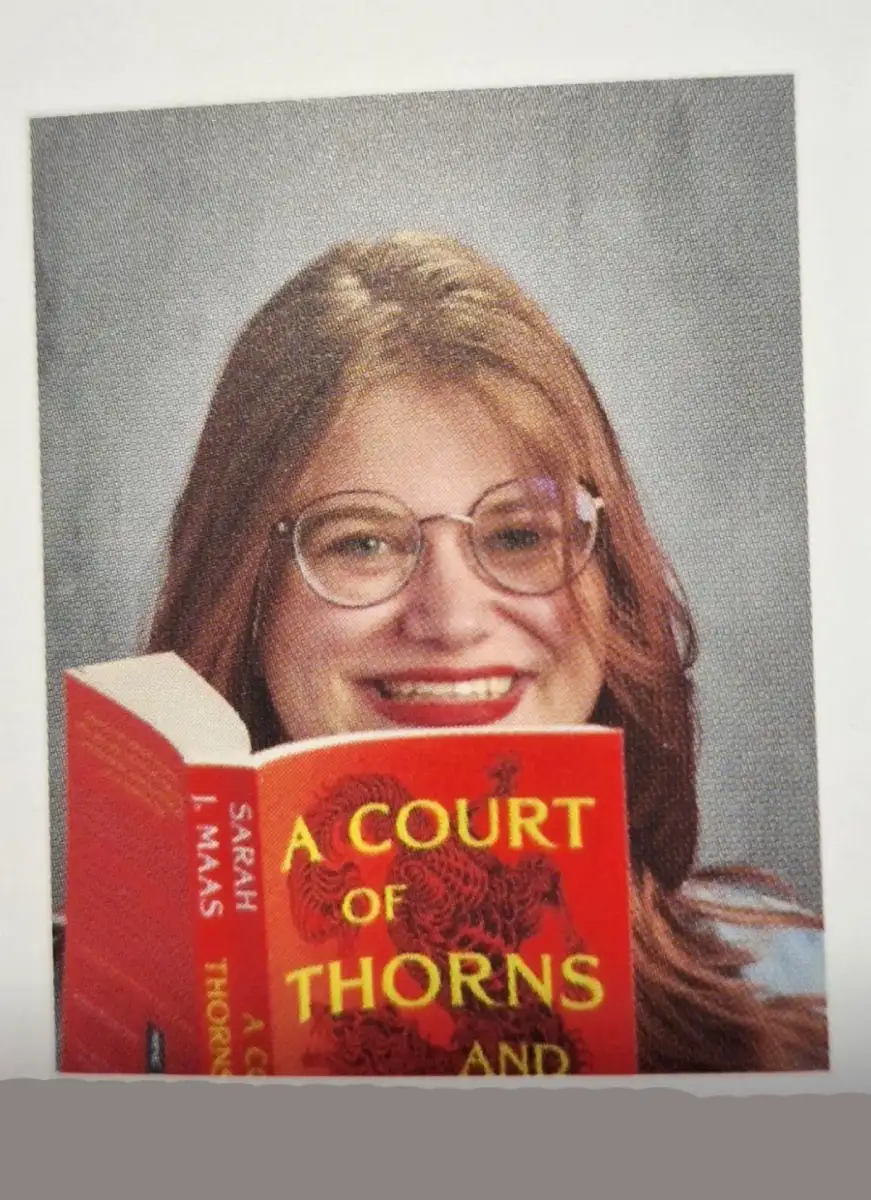 A person with long hair, glasses, and red lipstick smiles while holding up the book "A Court of Thorns and Roses" by Sarah J. Maas, partially covering their face. The background is a plain gray.
