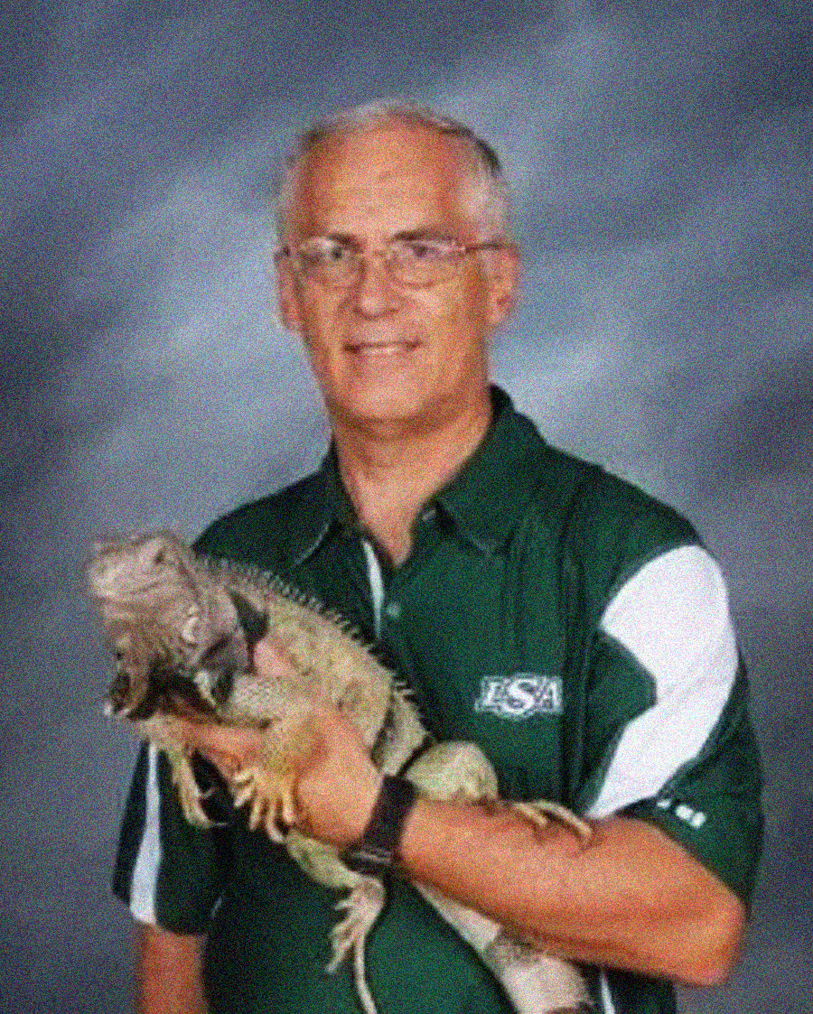 A man with short gray hair and glasses wearing a green and white polo shirt holds a large iguana. The background is gray and appears to be for a portrait photo.