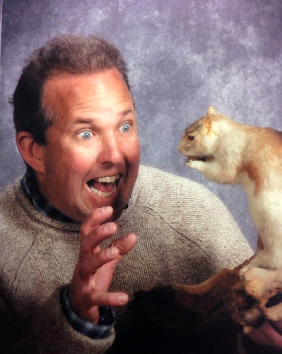 A man with an excited, surprised expression poses with a taxidermy squirrel, holding up his hand as if startled. He wears a beige sweater and plaid shirt, with a mottled gray background behind him.