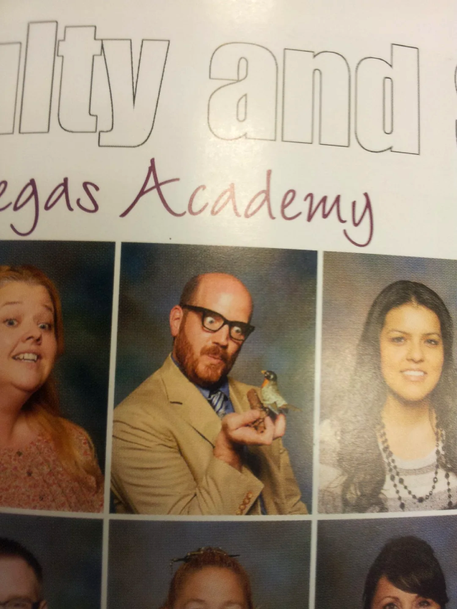 A man in a suit and glasses poses for a photo holding a small bird figurine, with two women’s portrait photos beside him on either side. The top of the image shows partial, cut-off text.