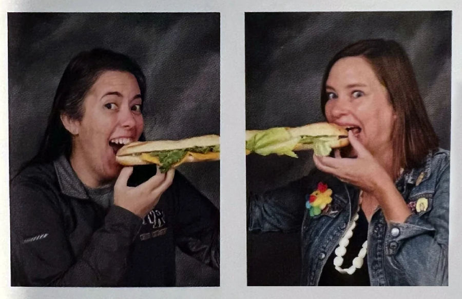 Two women face each other, both smiling and pretending to eat opposite ends of a long sandwich together, against a dark background.