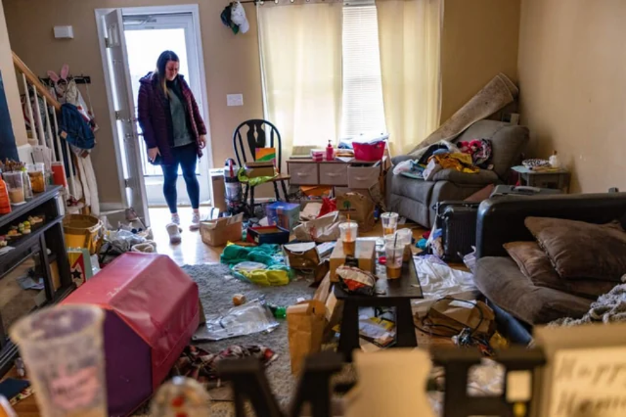 A woman stands in a cluttered, messy living room filled with trash, food containers, and scattered items on the floor and furniture, looking at the disarray with concern.