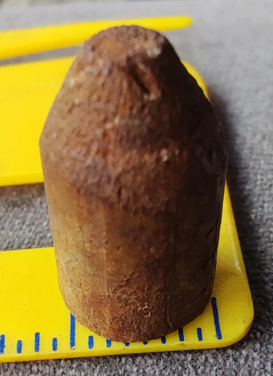 A rusty, bullet-shaped metal object sits upright on a yellow measuring tool, with blue markings visible. The object appears old and weathered.