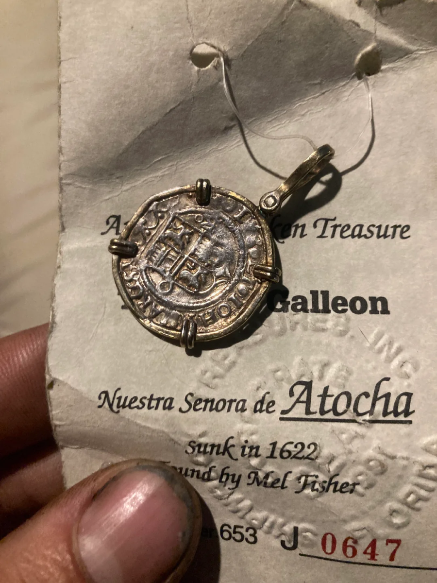 A hand holds a pendant made from a silver coin, attached to a card reading "Atocha, Authentic Sunken Treasure, Nuestra Senora de Atocha, Sunk in 1622, Found by Mel Fisher." The person's fingers appear dusty.